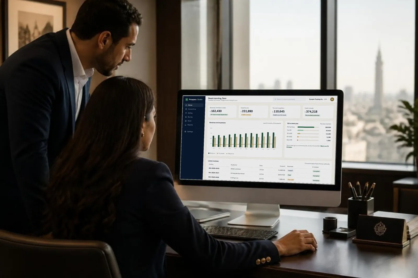 A finance manager and a colleague reviewing the Prospera Books dashboard on a desktop monitor in an office.